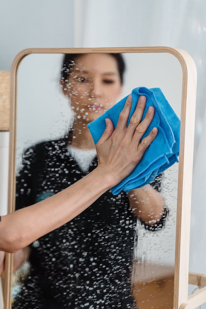 Close-up of a person wiping a mirror with a blue cloth, showing hands and reflection.